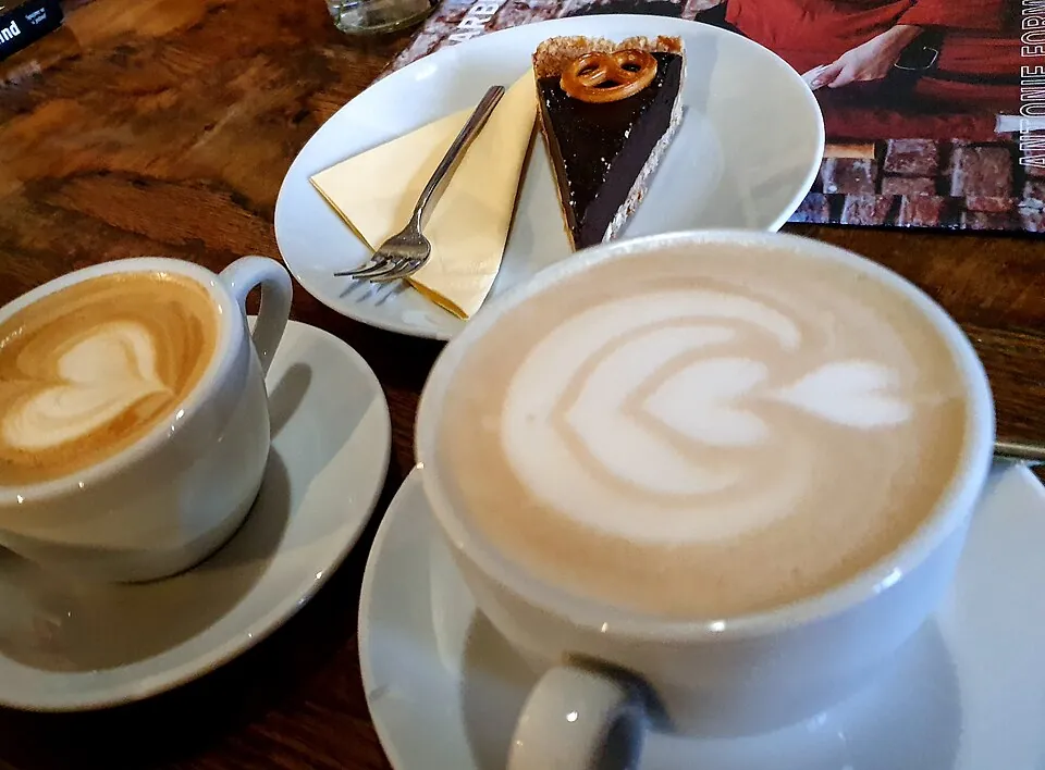 On the picture, we can see one chocolate cake with a small pretzel on top and two coffee cups with latte art (one heart and one flower with 